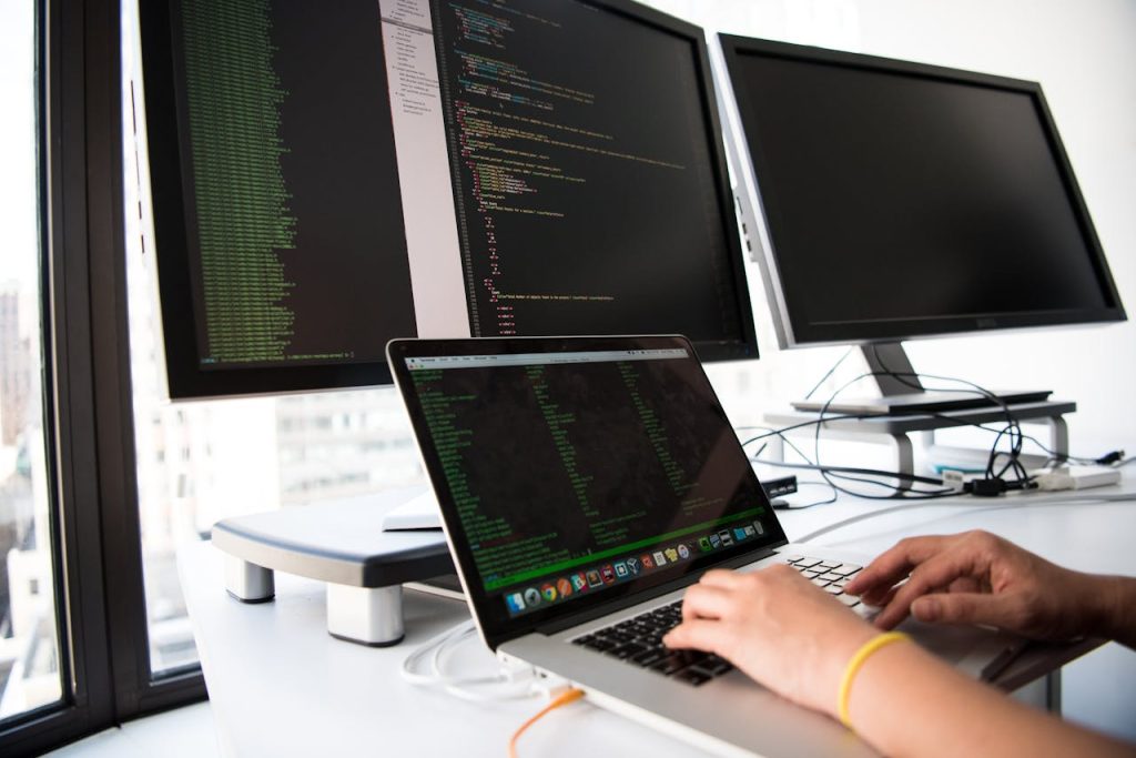 close-up-photo-of-person-typing-on-laptop-1181675 A developer writes code on a laptop in front of multiple monitors in an office setting.
